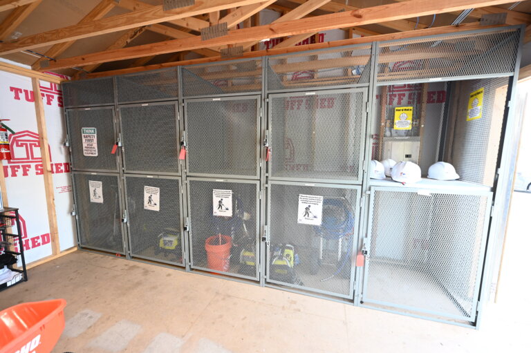 Metal storage cages with safety signs contain construction tools, buckets, and hard hats inside a partially built shed with exposed wooden beams and walls. The area is organized, with locked doors securing the equipment.