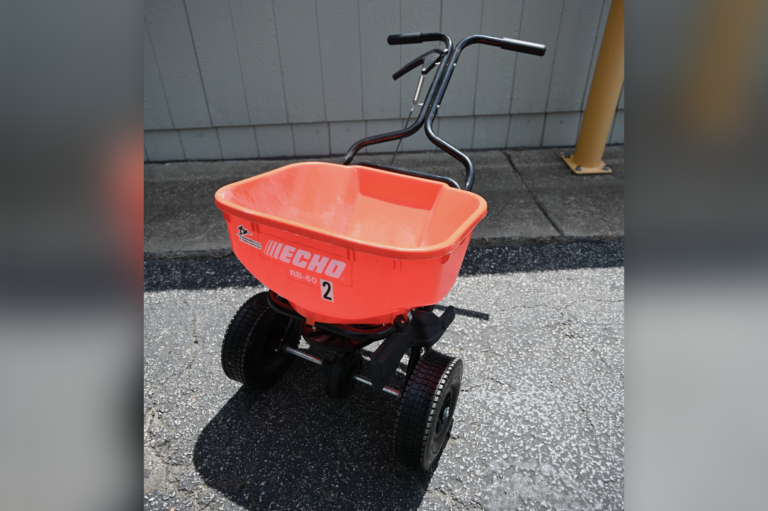 A bright orange Echo walk-behind broadcast spreader with black handles and wheels is placed on an outdoor concrete surface near a building.
