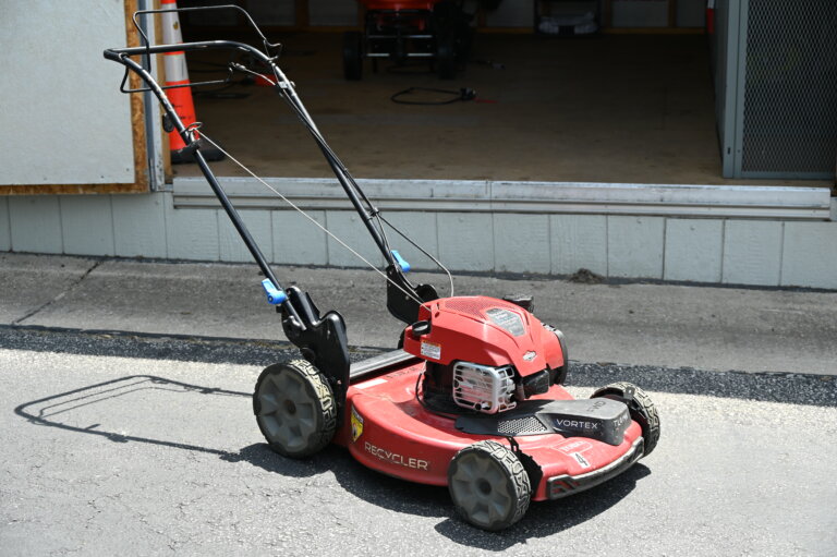 A red lawn mower with black handles is parked on a driveway in front of an open garage door.