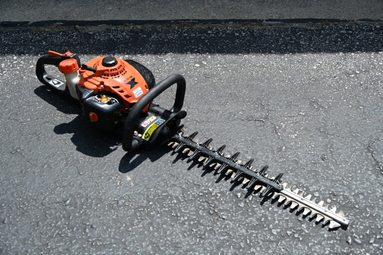 A gas-powered hedge trimmer with an orange engine and black blade is lying on an asphalt surface in sunlight.