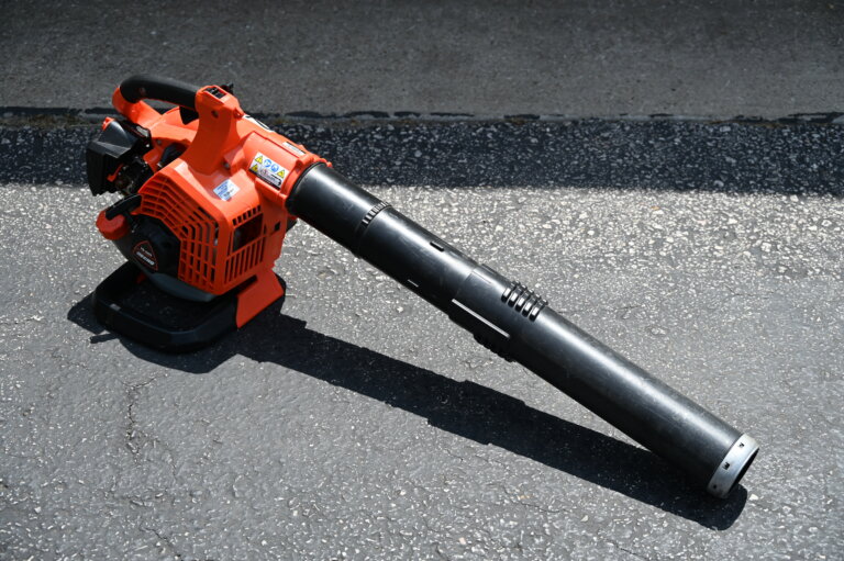 A red and black leaf blower lies on an asphalt surface, with sunlight casting shadows on the ground.