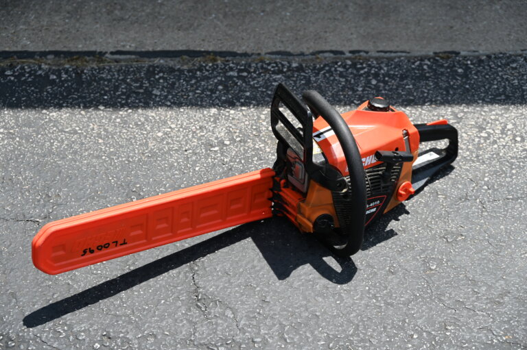 A red and black chainsaw with a protective cover on the blade rests on a sunlit asphalt surface.