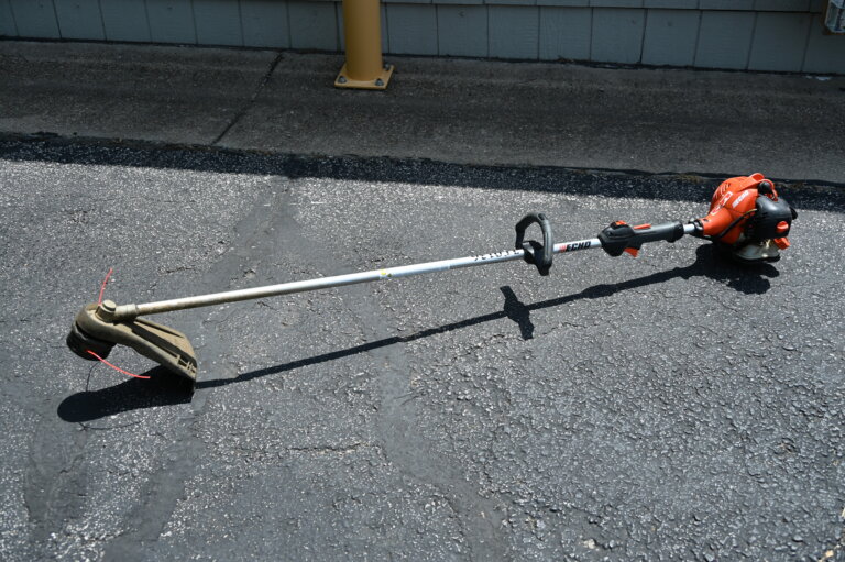 A gas-powered string trimmer with an orange engine lies on an asphalt surface near a building with a gray exterior wall.