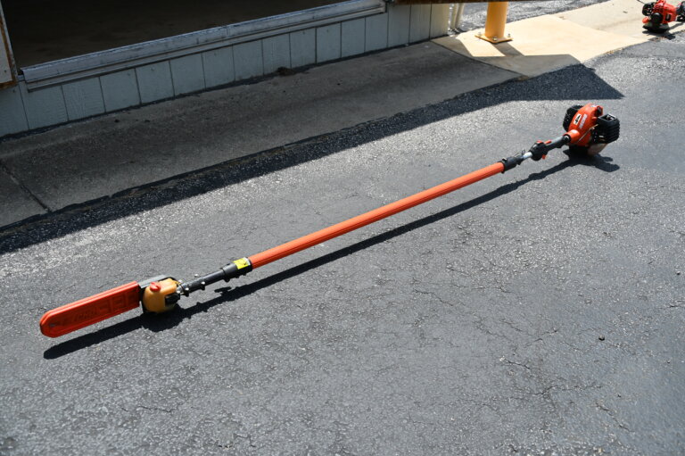 A long-handled orange pole saw with a chainsaw attachment lies on a paved surface outside a building, with sunlight casting shadows nearby.
