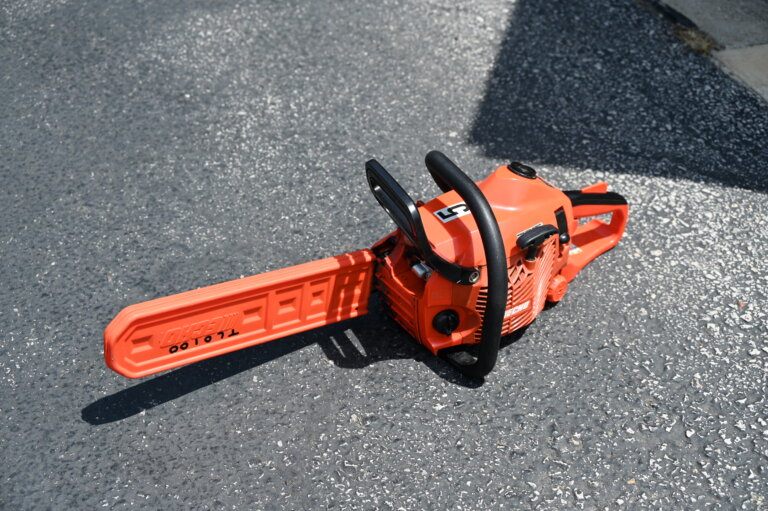 An orange chainsaw with a protective blade cover lies on a textured asphalt surface, partially in sunlight and partially in shade.