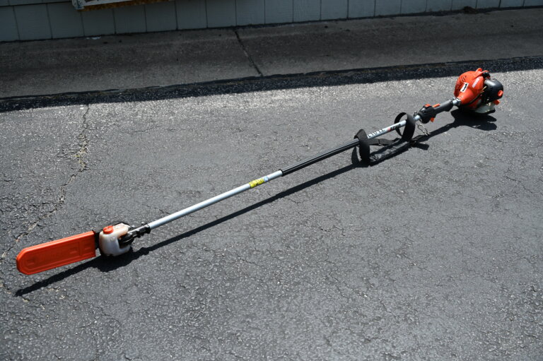 A long-handled pole saw with an orange blade cover is lying on a blacktop driveway near a curb and a building wall.
