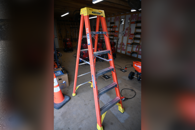 A red and yellow step ladder stands open inside a garage or workshop, surrounded by various tools, equipment, and a traffic cone on the concrete floor.