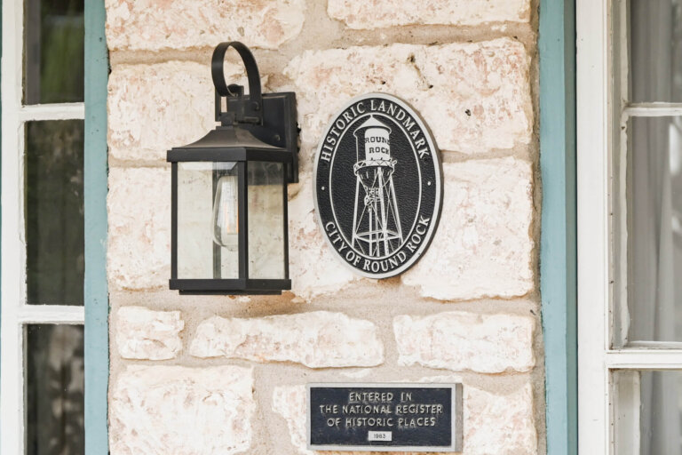 A stone wall displays a lantern, a round Historic Landmark City of Round Rock plaque, and a rectangular plaque noting entry in the National Register of Historic Places in 1983.