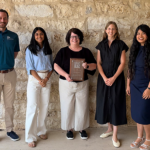Five people stand smiling in front of a stone wall; the person in the middle holds a plaque. The group is dressed in business casual attire, and they appear to be at an award or recognition event.