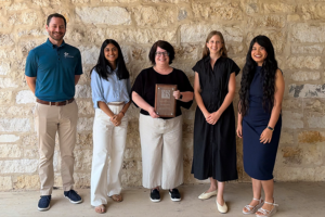 Five people stand smiling in front of a stone wall; the person in the middle holds a plaque. The group is dressed in business casual attire, and they appear to be at an award or recognition event.