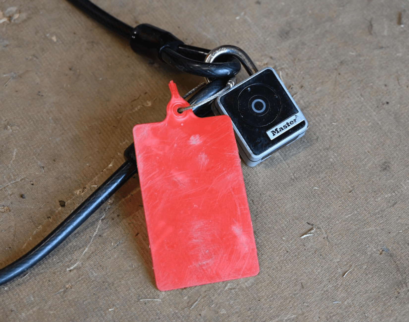 A close-up of a black cable secured by a Master padlock with a blank, scratched red plastic tag attached, all resting on a textured brown surface.