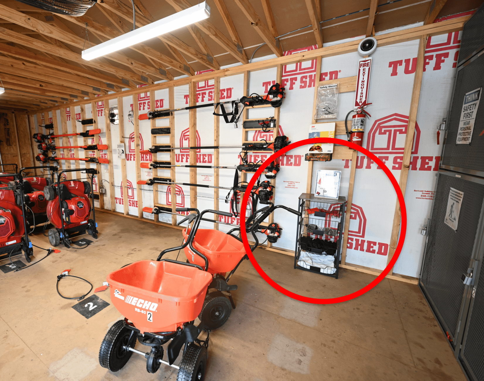 A shed interior with organized landscaping tools on the walls, red equipment in the foreground, and a red circle highlighting a rack of safety helmets, goggles, and ear protection gear against the wall on the right.