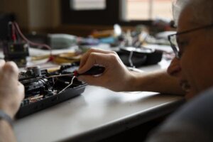 A person uses a screwdriver to repair or work on an electronic device at a table, with tools and components scattered in the background.