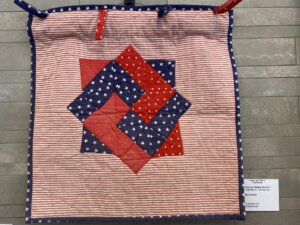 A small quilt with red, white, and blue star and stripe patterns arranged in a geometric design, displayed on a gray surface with an information card visible in the bottom right corner.