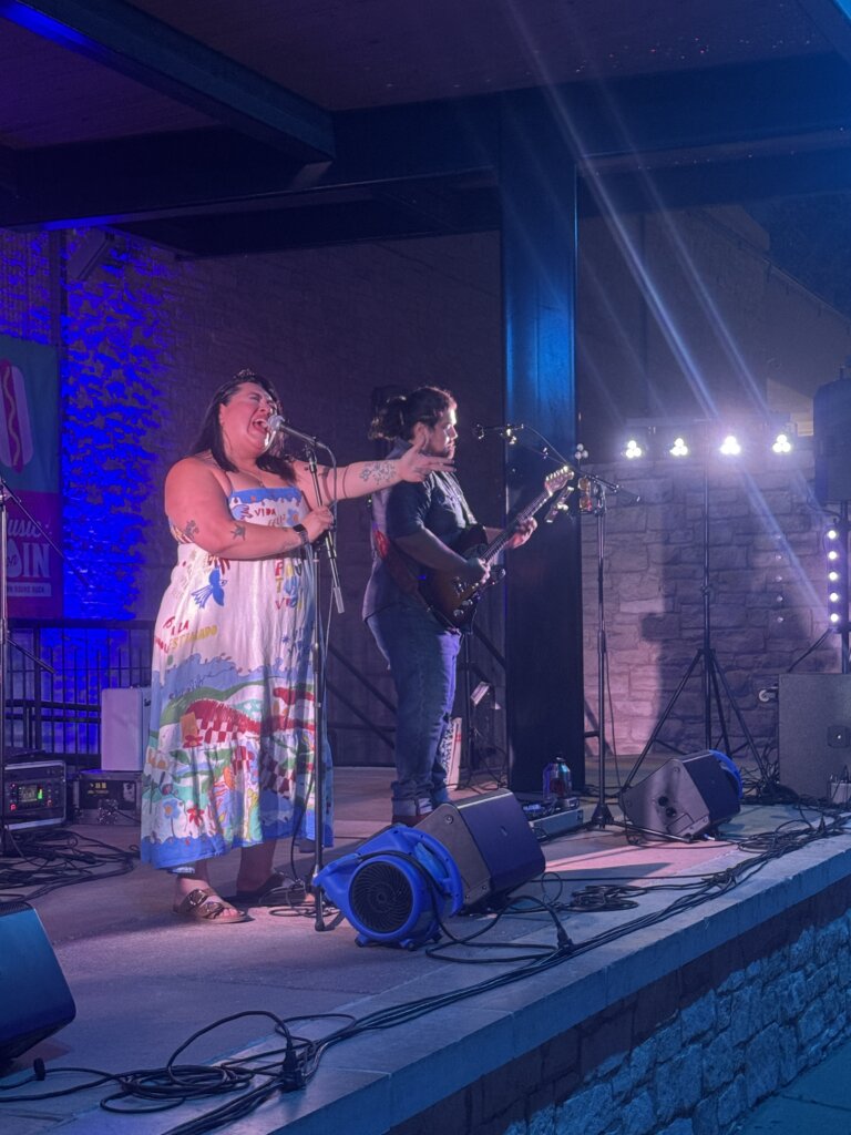 A woman in a colorful dress sings on stage with a microphone while a guitarist plays beside her under blue and purple lighting.