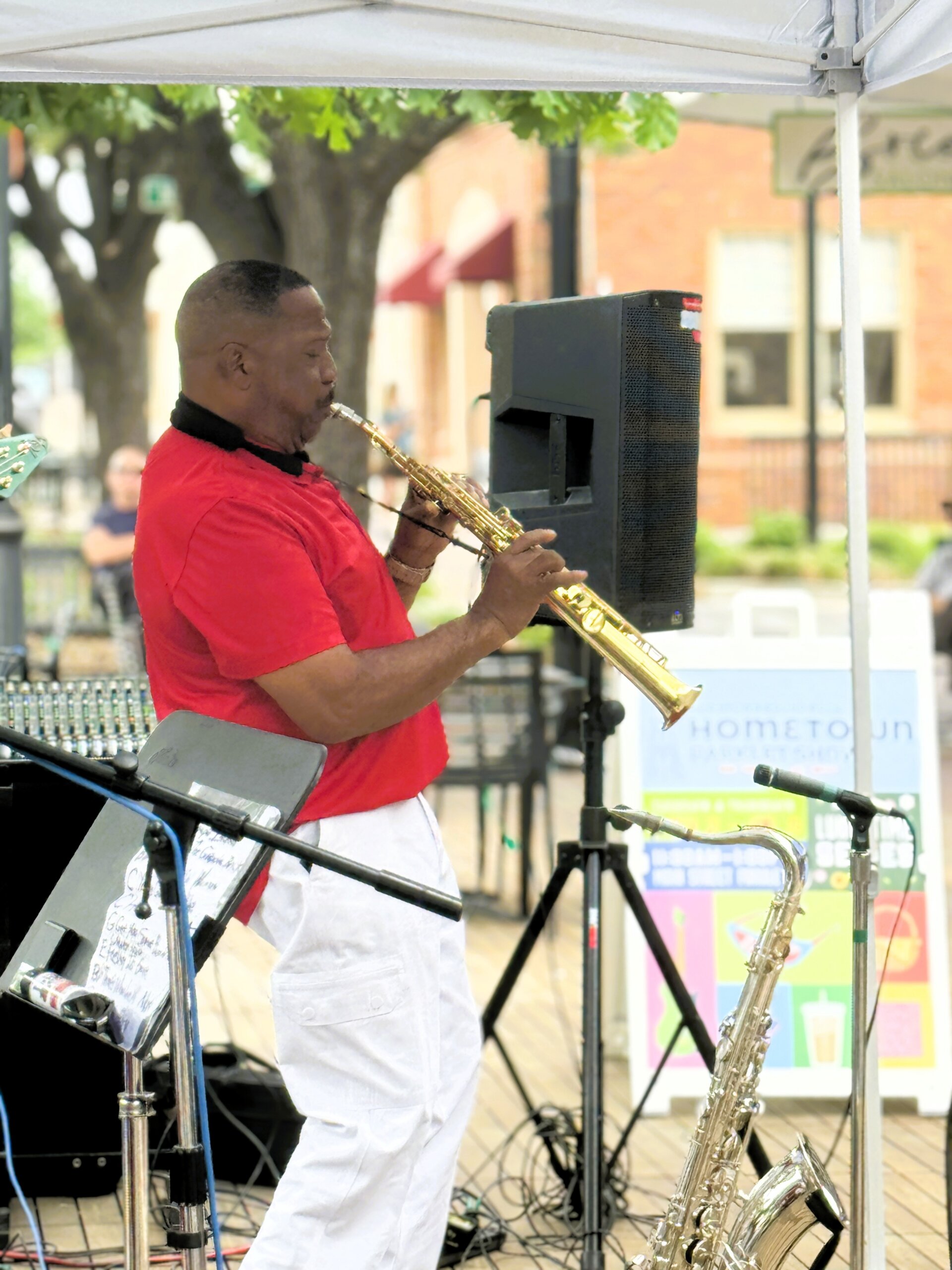 A man in a red shirt and white pants plays a saxophone at an outdoor event. Other instruments and equipment are nearby, with trees and buildings in the background.