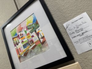 Framed colorful watercolor painting of a street with Main St. Round Rock sign, next to a gallery label listing artist Judy Blund, artwork title, and medium as watercolor and pencil.
