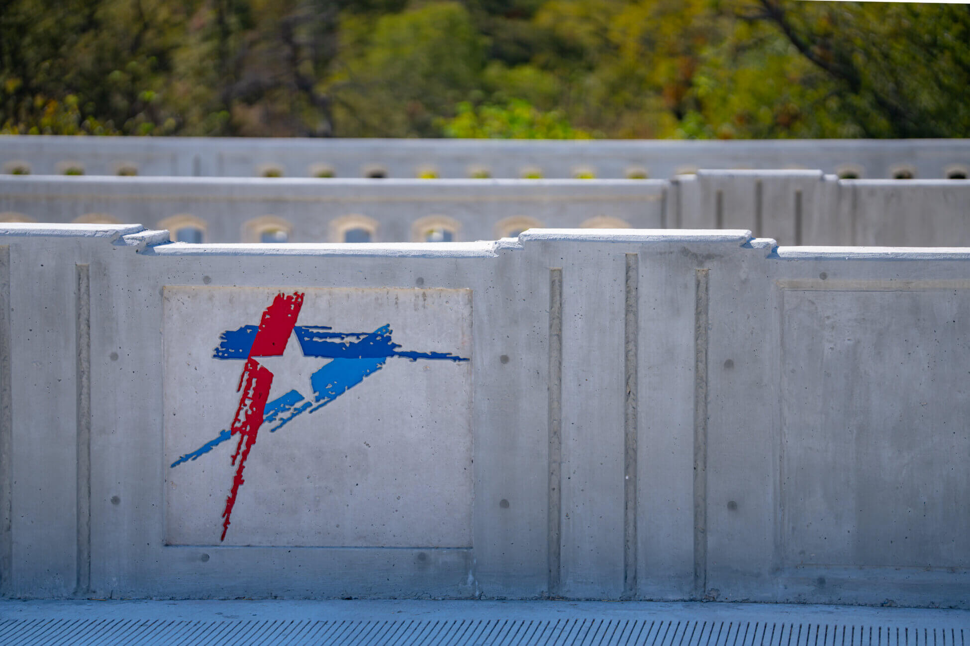 Concrete barrier with a colorful abstract design in red and blue, featuring a star shape. Trees and greenery are visible in the blurred background beyond the wall.