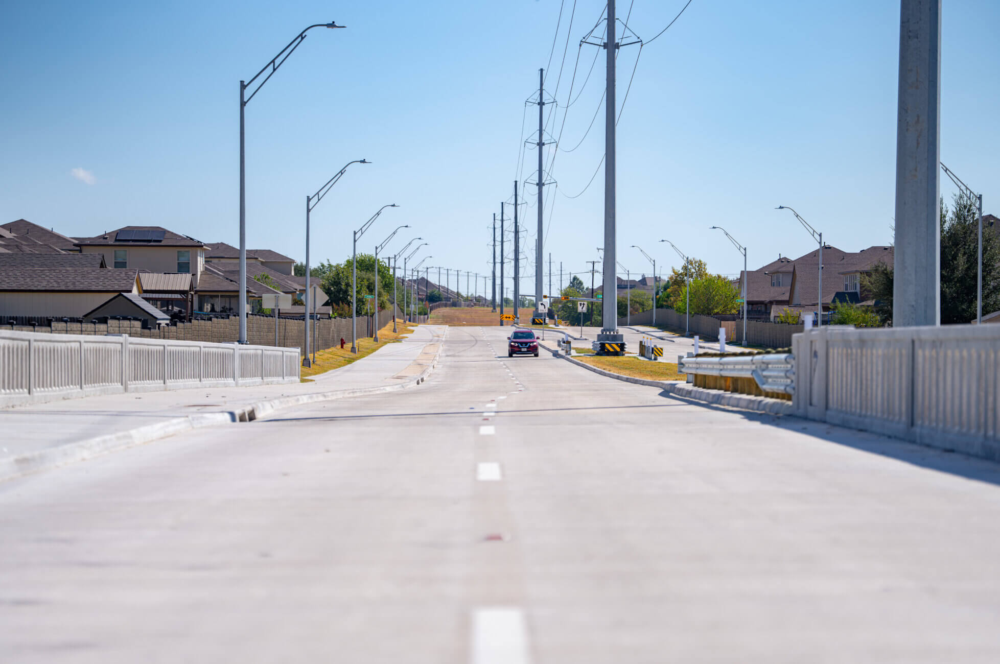 A wide, empty concrete road with one car driving away, lined with streetlights and power lines, houses on both sides, under a clear blue sky.