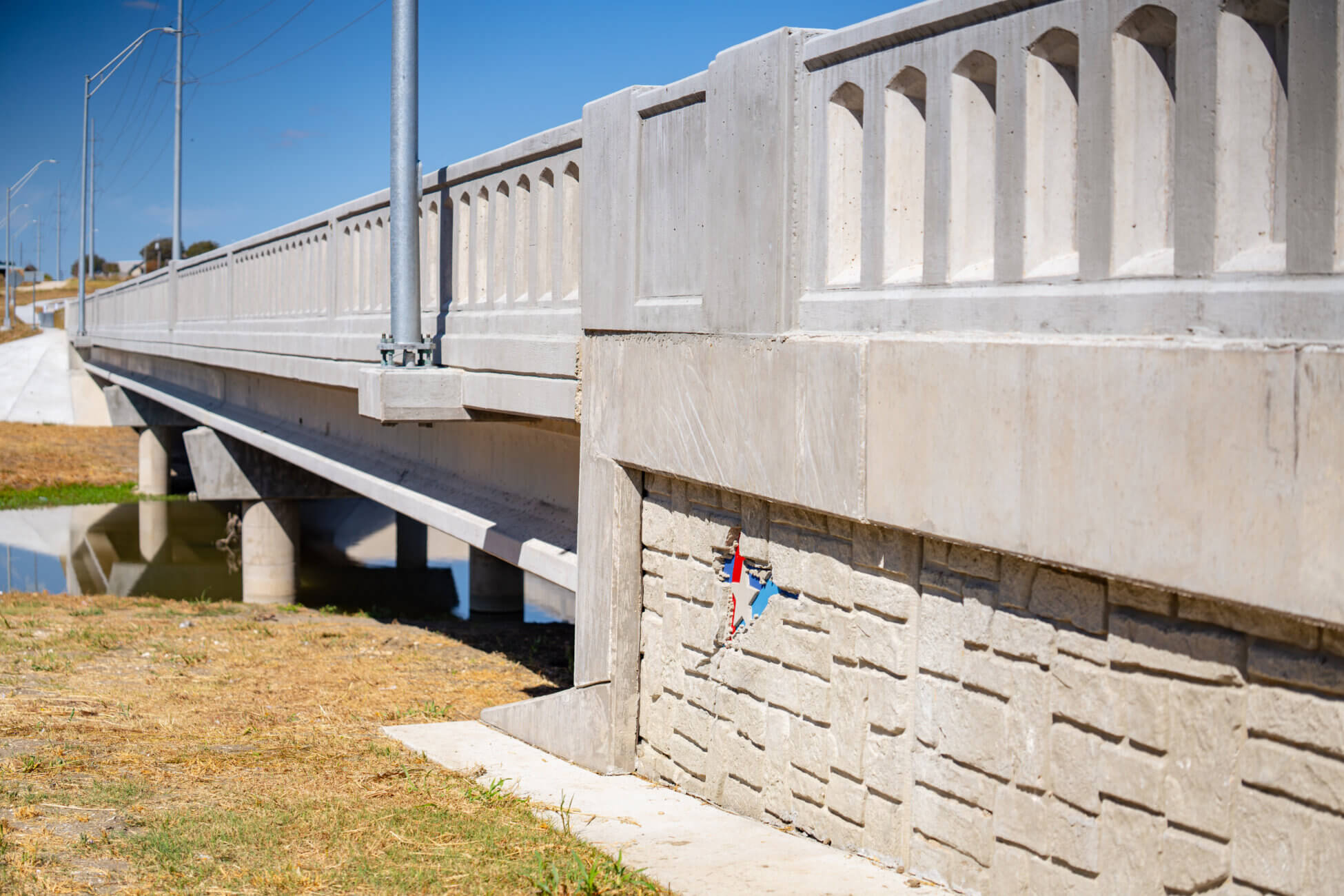 A concrete bridge with decorative railings crosses over a small body of water, with dry grass and a paved embankment visible beneath and beside the structure.