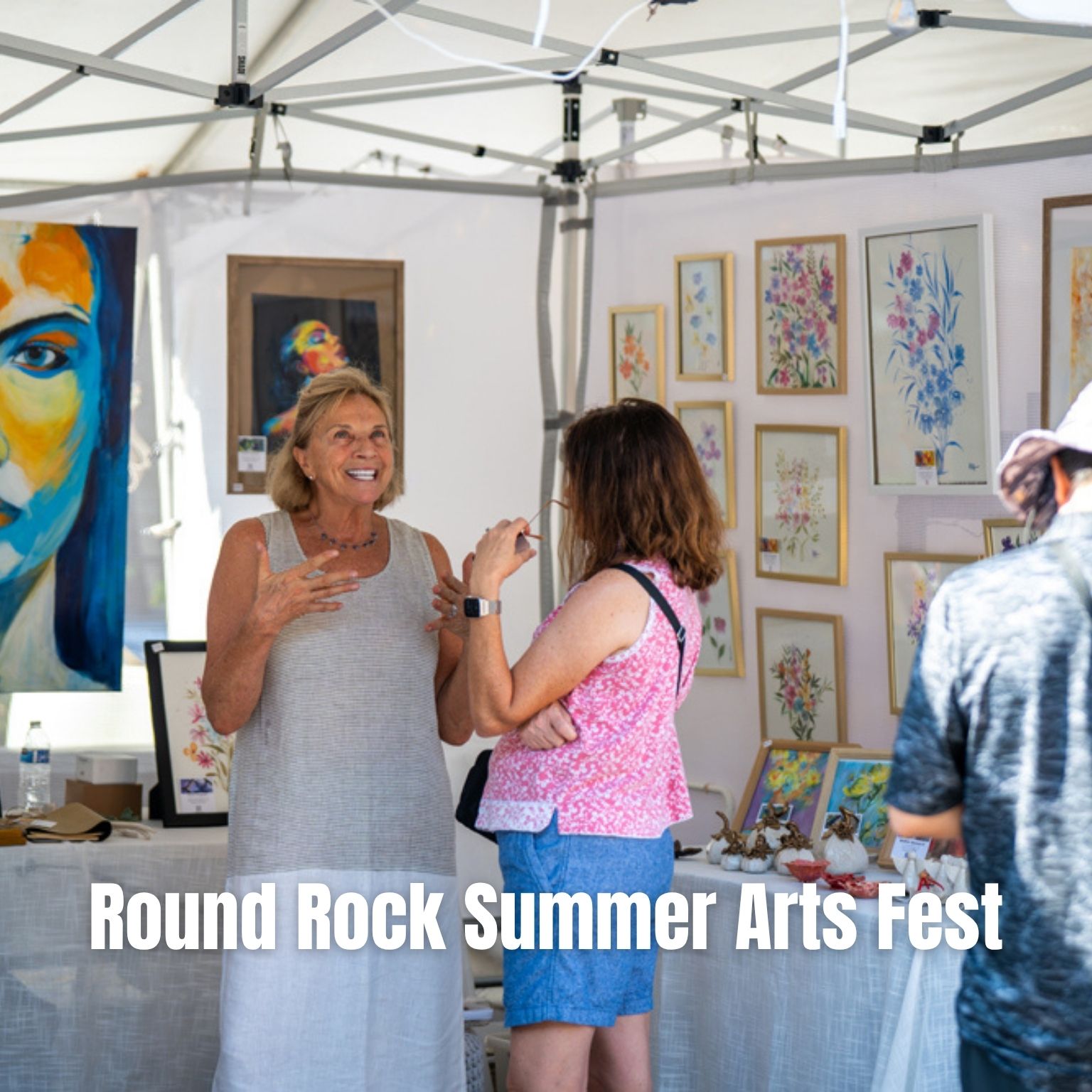 Two women chat in front of art displays under a white tent at an outdoor arts festival. Colorful paintings hang and sit on tables. Text reads: Round Rock Summer Arts Fest..
