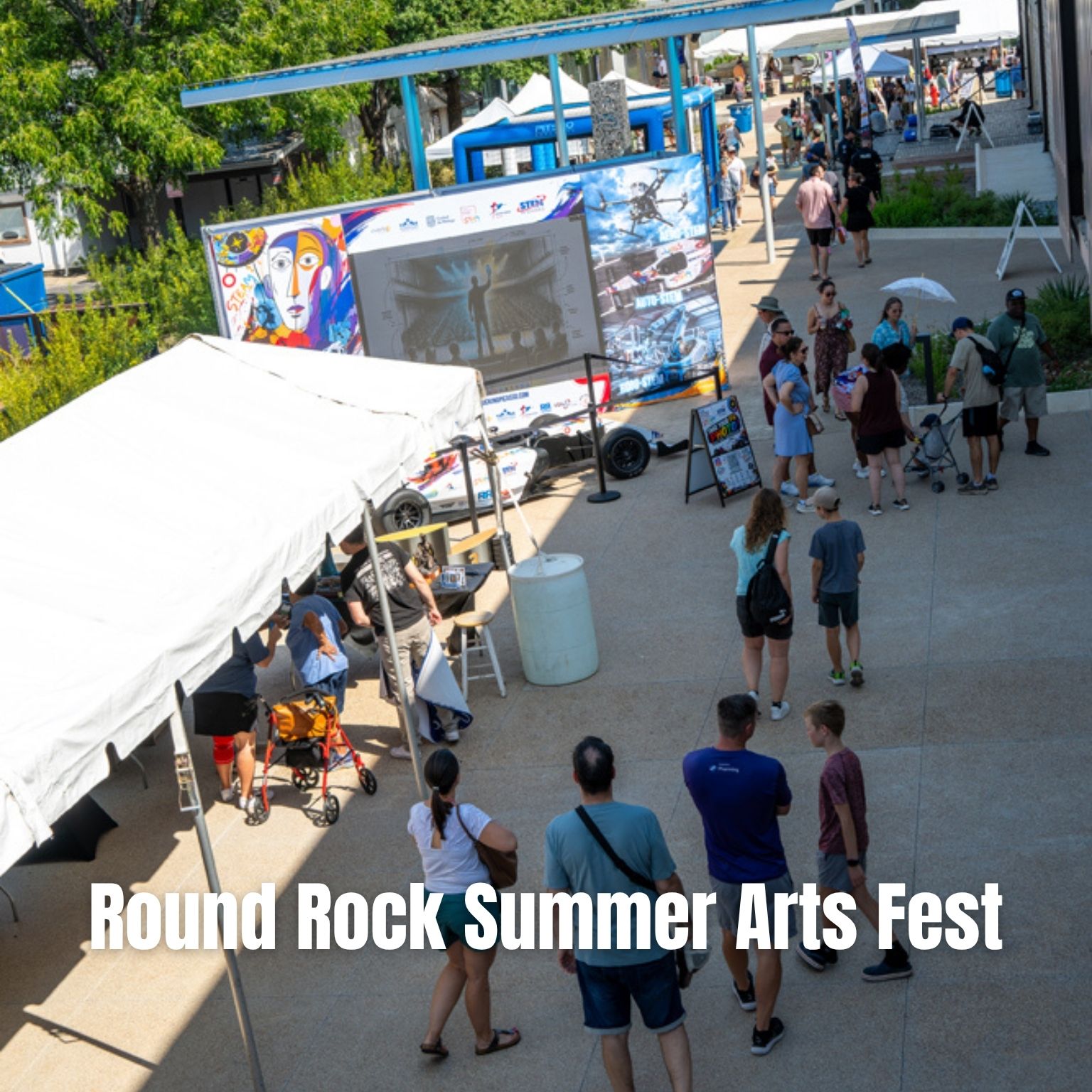 People attend the Round Rock Summer Arts Fest outdoors, walking past tents and an art display with a large screen. Bright sunny weather, attendees of all ages, and art banners are visible.