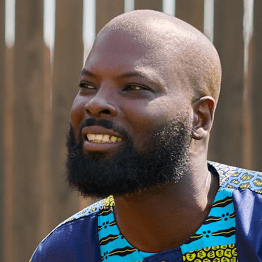A man with a full beard and shaved head smiles while wearing a colorful patterned shirt. A wooden fence is visible in the blurred background.