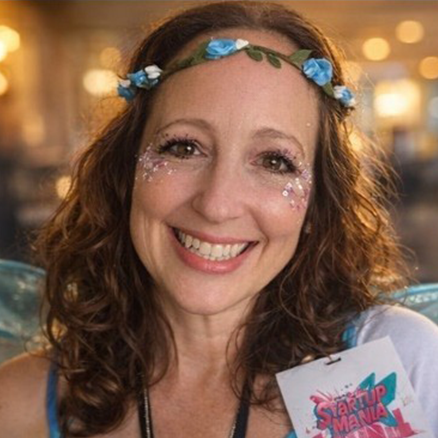 Smiling woman with curly hair wears a blue flower crown and sparkly makeup, holding a Startup Mania badge. The background is softly blurred with warm lighting.
