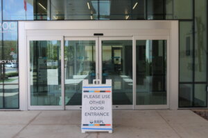 A white sign in front of closed glass doors reads, PLEASE USE OTHER DOOR ENTRANCE. The entrance appears to be at a public building.