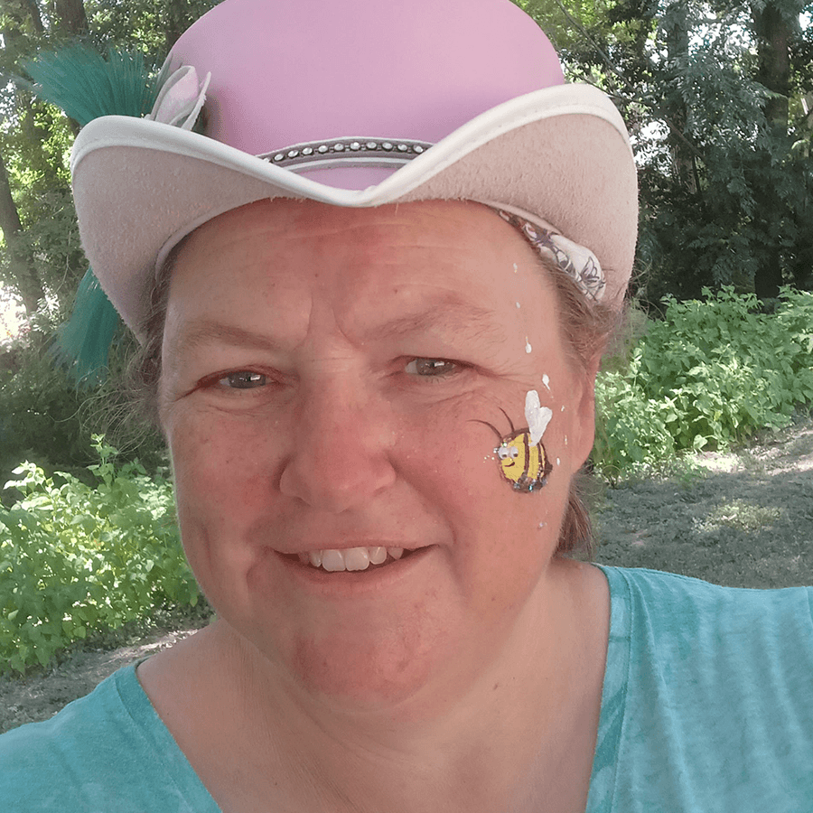Smiling person in a light pink cowboy hat with a painted bee on their cheek, standing outdoors in front of green plants and trees.