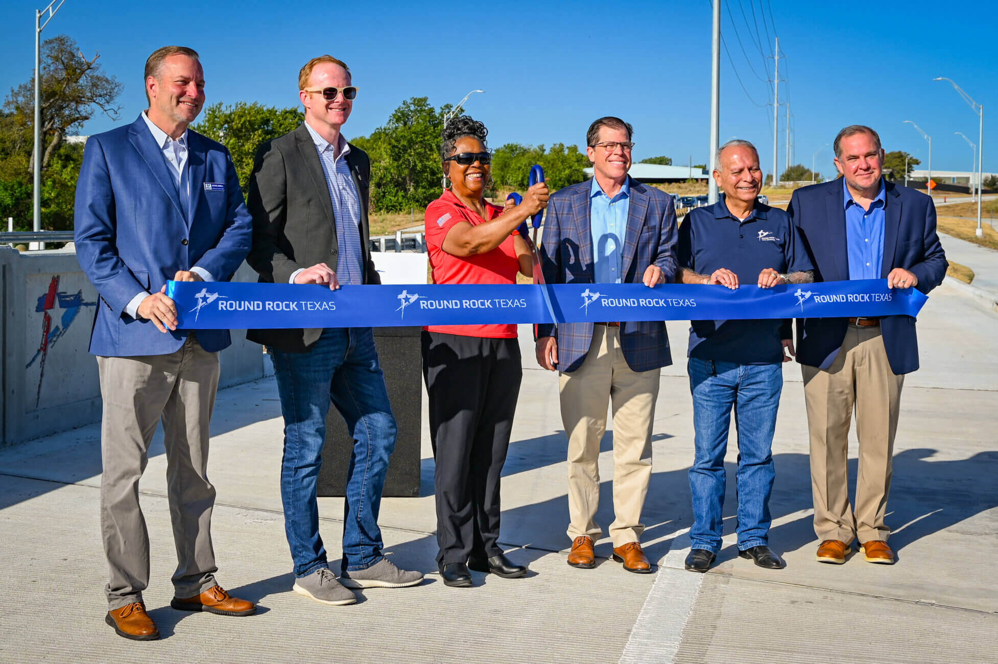 Six people stand on a new roadway, smiling and holding a blue ribbon that says Round Rock Texas. One woman holds large scissors, suggesting a ribbon-cutting ceremony. Blue sky and trees are in the background.