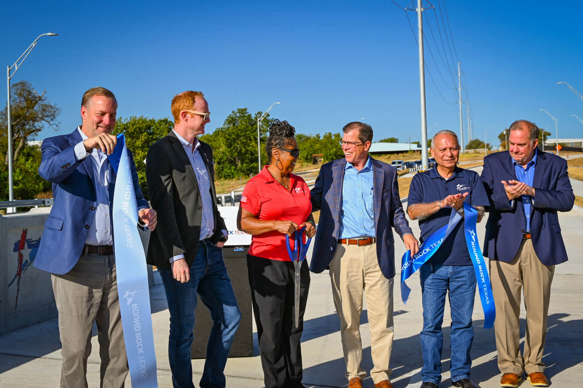 Six people stand on a new bridge, smiling and holding a blue ribbon at a ribbon-cutting ceremony under a clear blue sky.