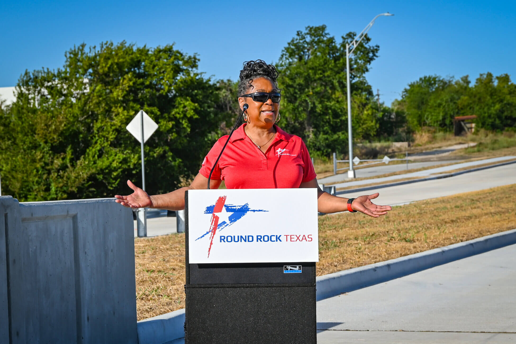 A woman wearing sunglasses and a red polo shirt stands at a podium labeled Round Rock Texas, speaking outdoors on a sunny day with roads and greenery in the background.