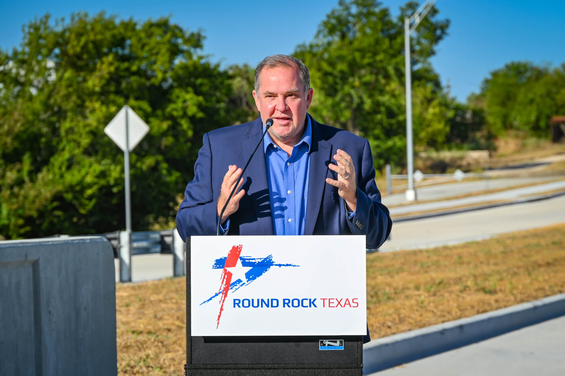 A man in a blue shirt speaks at a podium with a Round Rock Texas sign outdoors. Trees and a road are visible in the background.