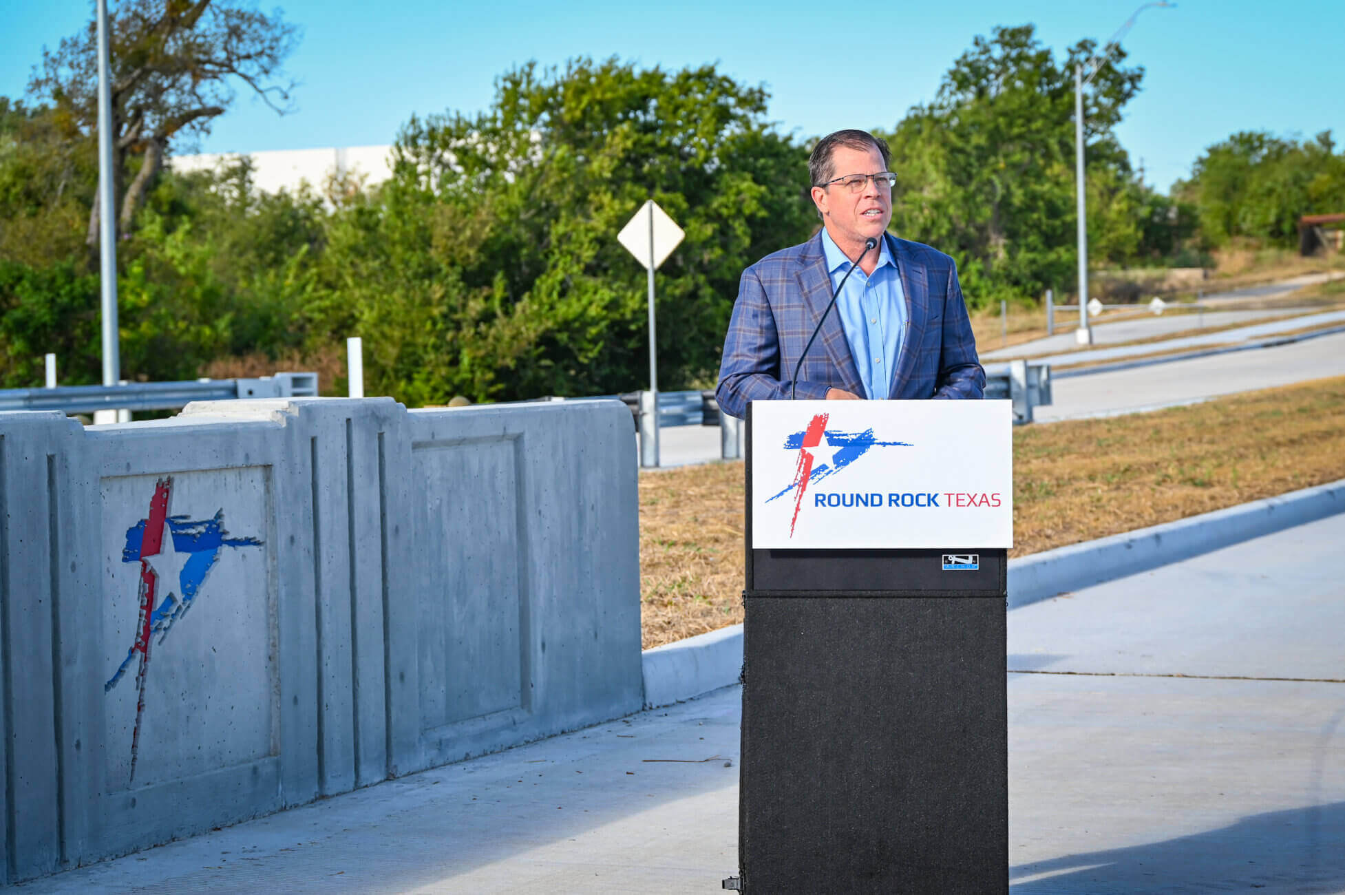 A man in a blue suit speaks at a podium with a Round Rock Texas logo, outdoors near a newly constructed road with concrete barriers and trees in the background.