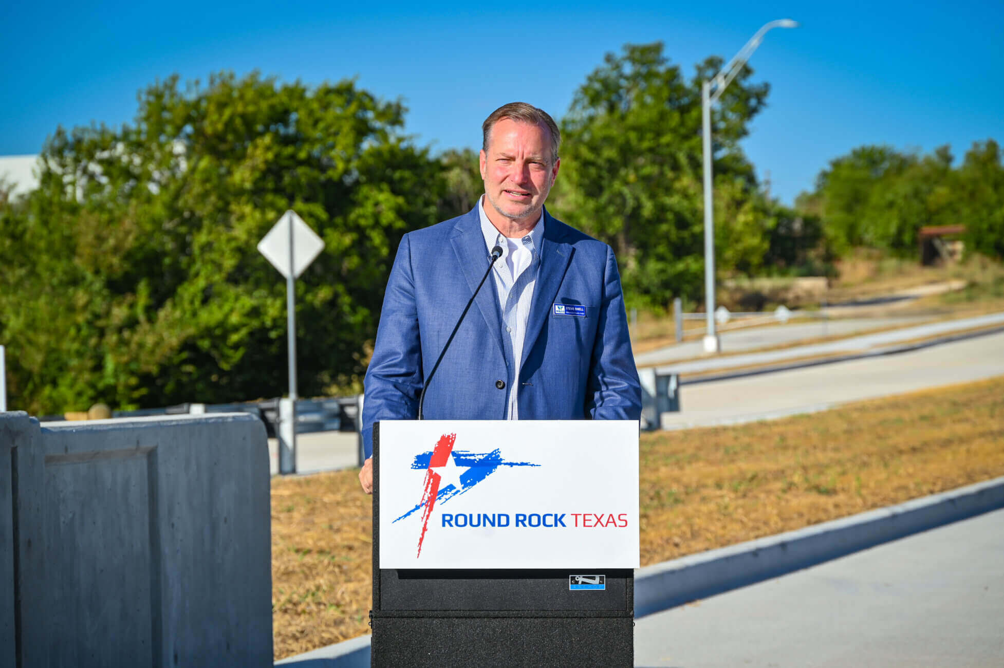 A man in a blue suit stands at a podium labeled Round Rock Texas, speaking outdoors on a sunny day with trees and a road in the background.