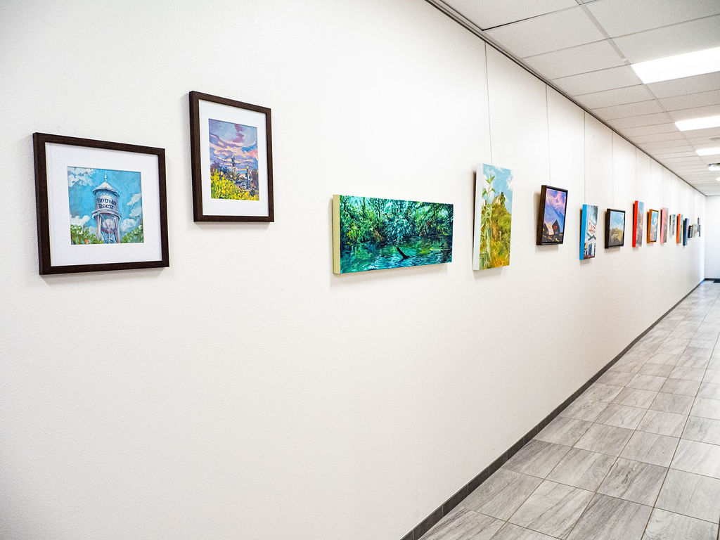 A hallway with a tiled floor features several colorful paintings and framed artworks hanging on a white wall, reminiscent of pieces you might find at music and arts round rock. The corridor stretches into the distance, with a ceiling of white panels and fluorescent lighting.