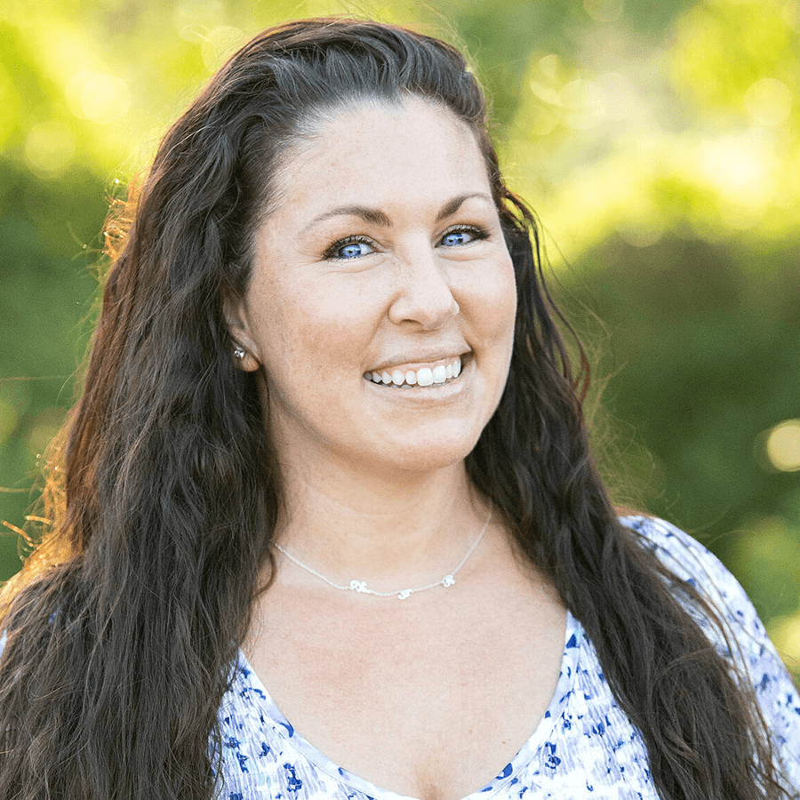 A woman with long, wavy brown hair smiles outdoors. She is wearing a white and blue patterned top and a delicate necklace. The background is blurred greenery with sunlight filtering through.