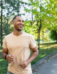 A smiling man in a light t-shirt jogs along a sunlit park path with green trees in the background, enjoying the scenic route promoted by the Round Rock Trails Plan.