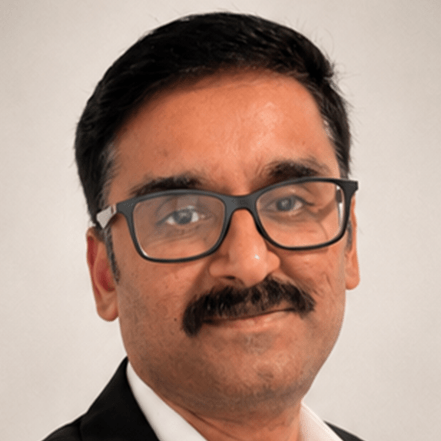 A man with short black hair, glasses, and a mustache is smiling slightly. He is wearing a white shirt and a dark jacket. The background is plain and light-colored.