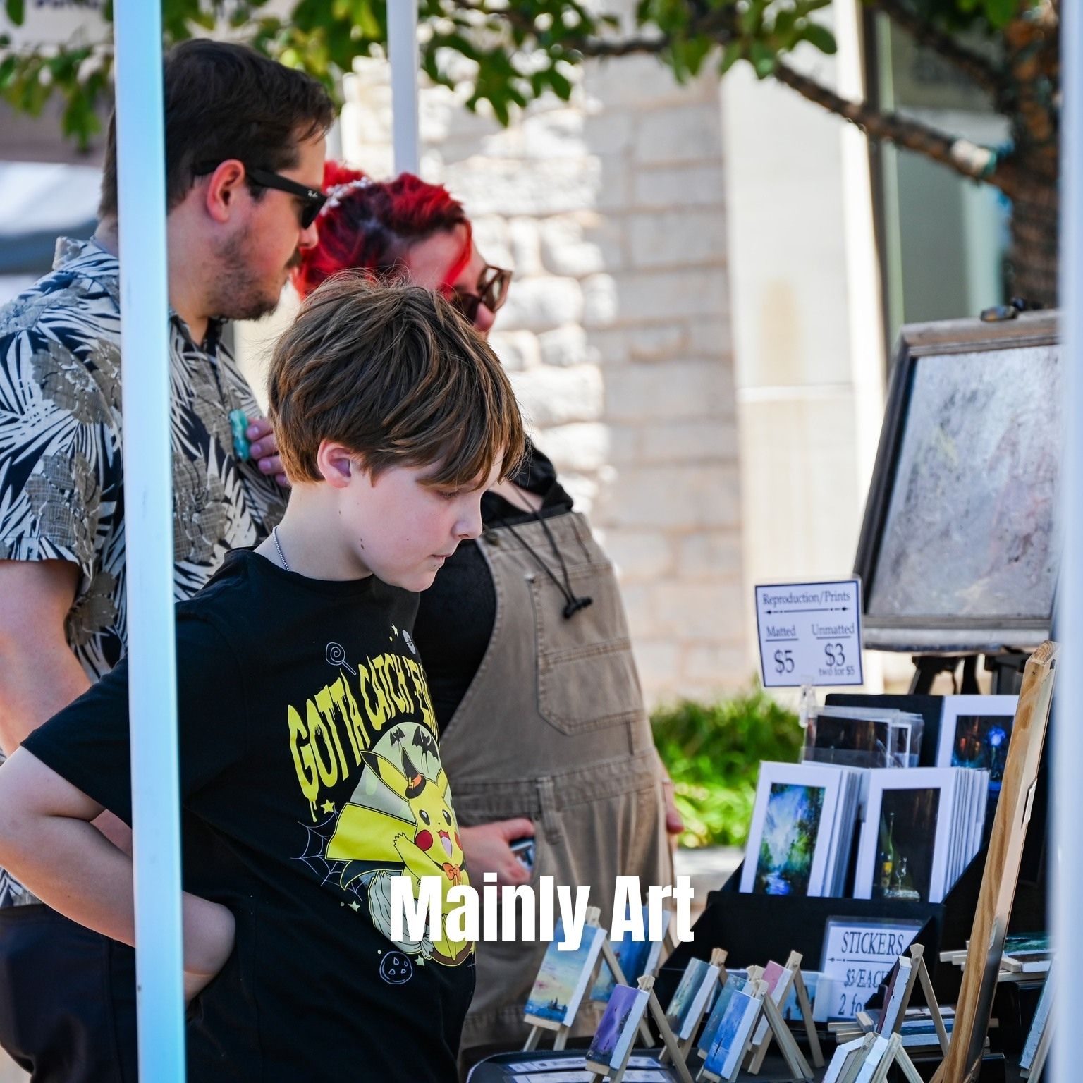 A child and two adults browse art prints and stickers at an outdoor market booth. The child wears a Pikachu T-shirt. Art displays and price signs are visible on the table, with trees and a stone wall in the background.
