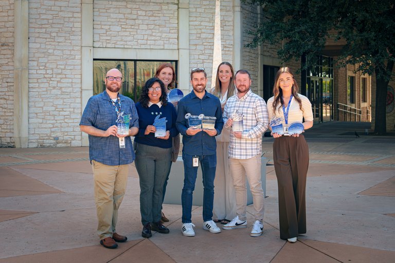 Seven smiling professionals pose outdoors holding awards in front of a stone building.