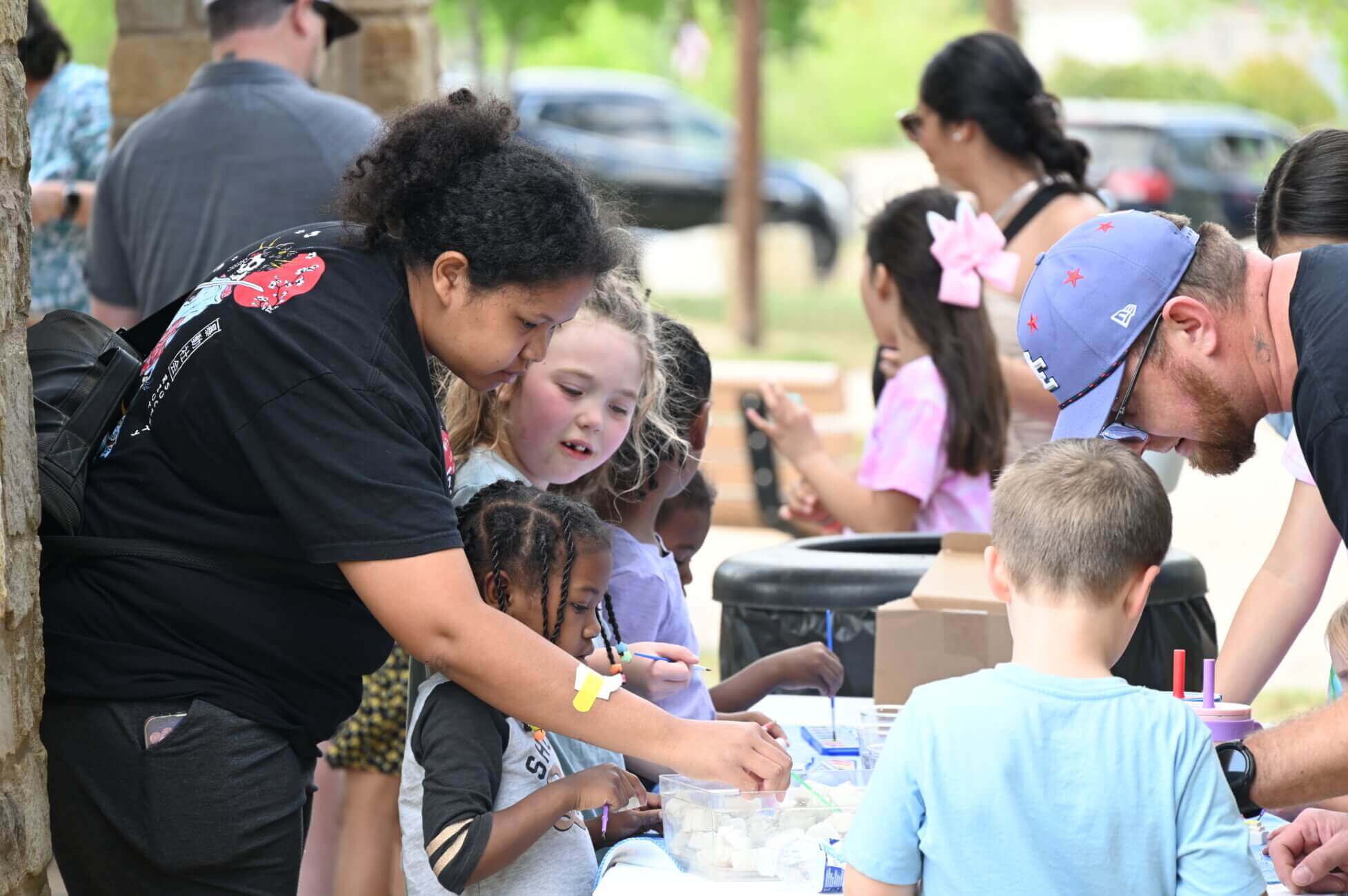 A woman and a man help a group of children participate in an outdoor craft activity at a table.