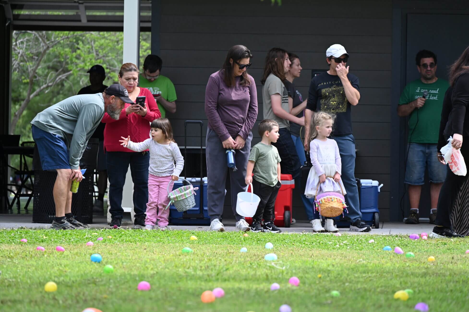 A group of children and adults standing on grass filled with scattered colorful plastic Easter eggs.