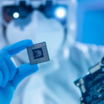 A person in protective gear and blue gloves holds a computer processor chip in front of a circuit board.