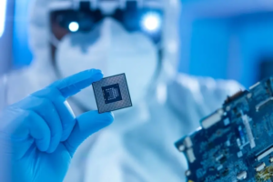 A person in protective gear and blue gloves holds a computer processor chip in front of a circuit board.