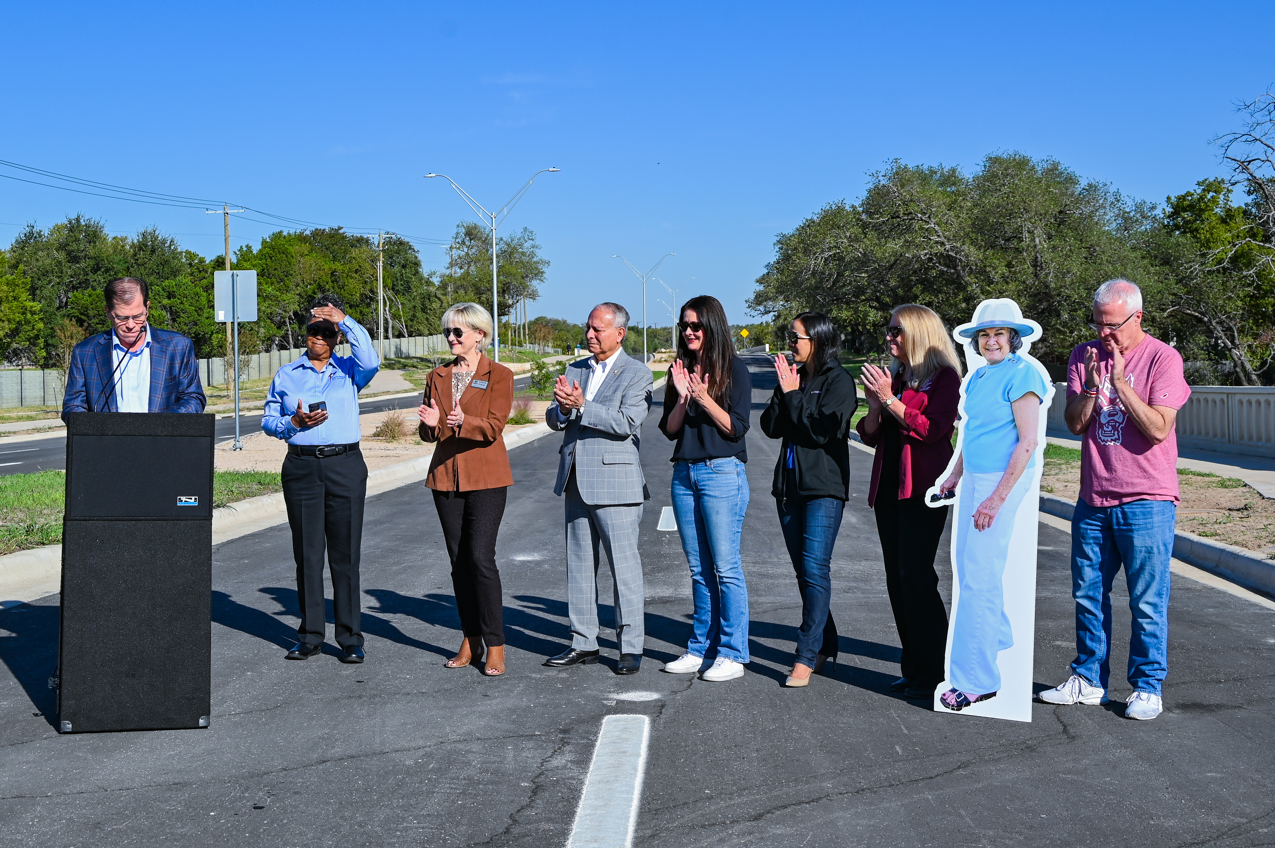 A man speaks at a podium as a group of people and a cardboard cutout of a woman stand on a paved road and applaud.