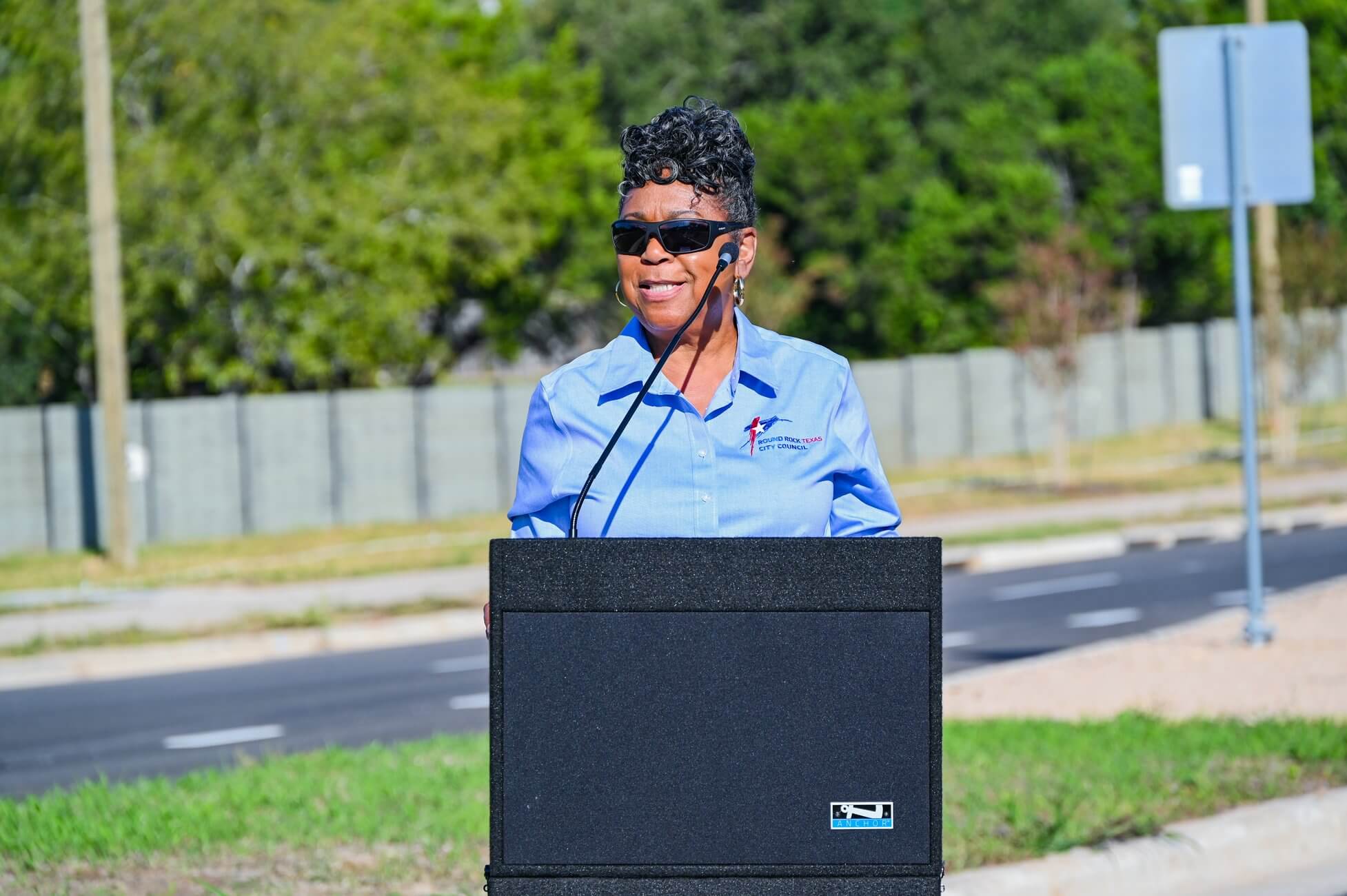 A woman wearing sunglasses and a blue shirt speaks outside at a podium on a sunny day, with trees, a road, and a fence in the background.