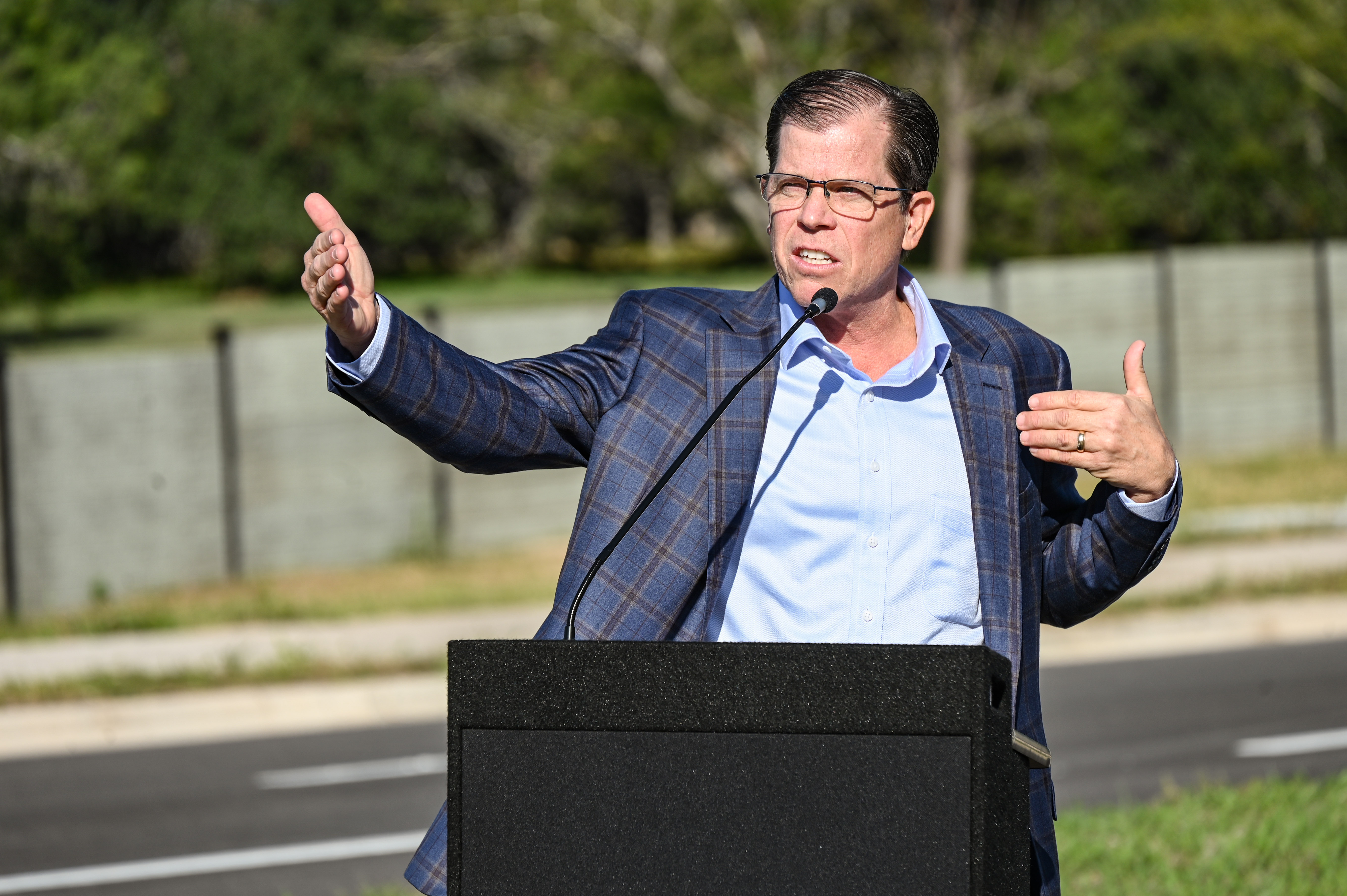 Marty Walsh wearing a blue plaid suit jacket and light blue dress shirt, gesturing while speaking at a podium outdoors.