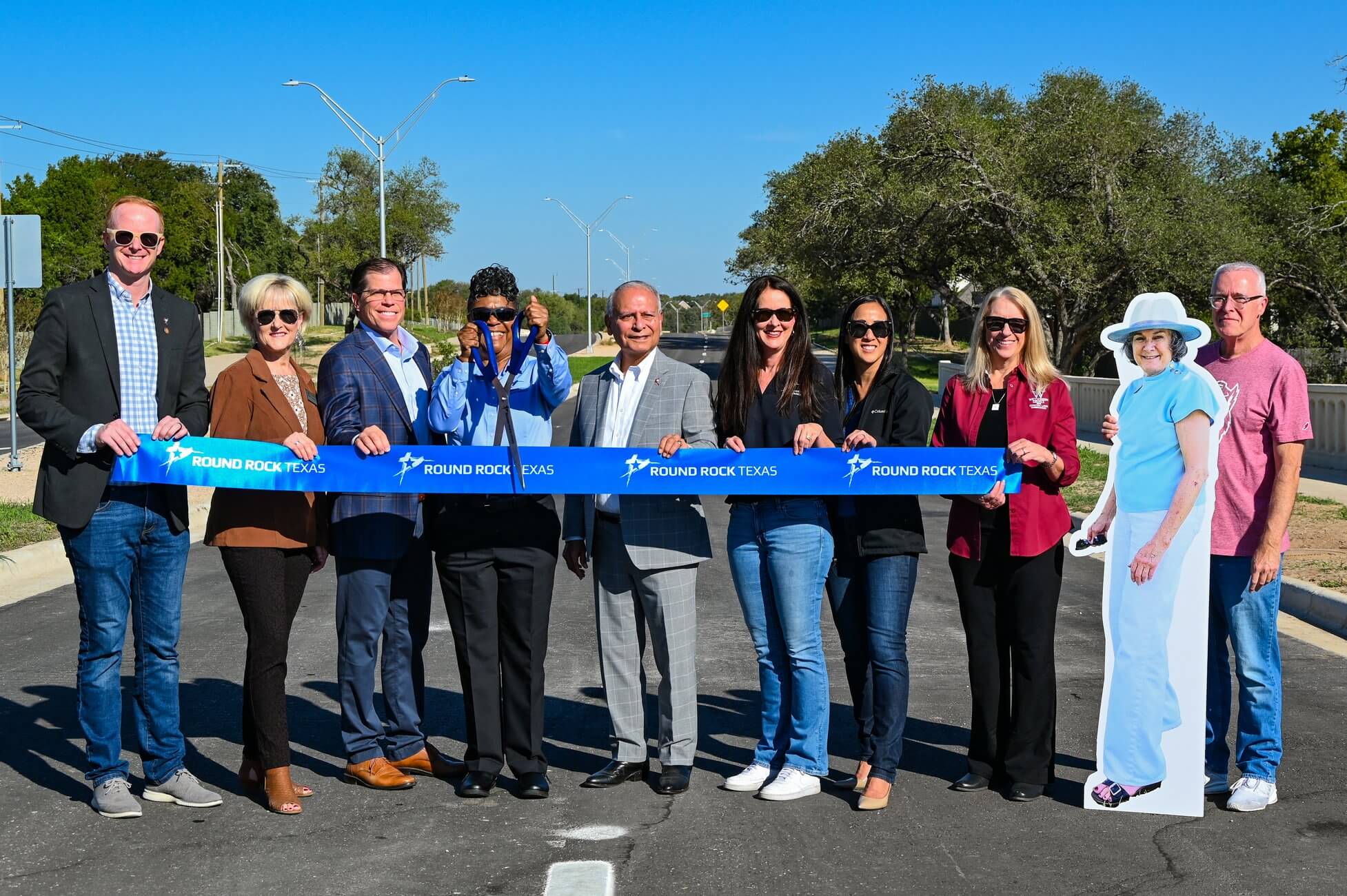 A group of people standing on a new road holding a blue ribbon for a ribbon-cutting ceremony, with a life-sized cutout of a woman standing on the right.